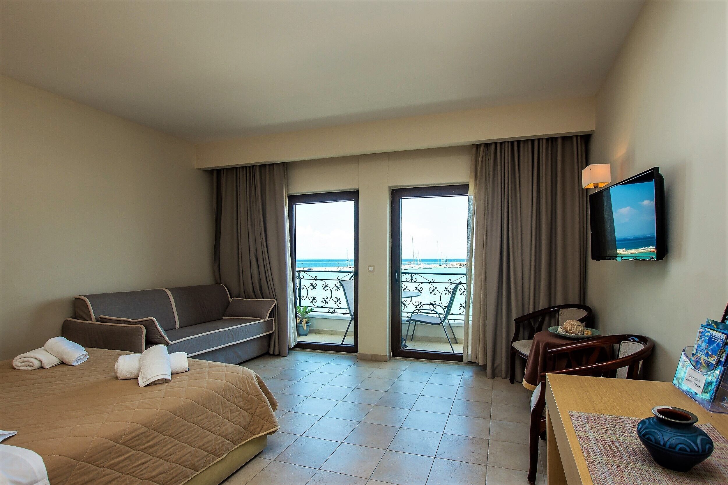 A hotel room with a bed, a sofa, a dining table, and a TV. There is a balcony with a view of the sea.