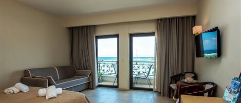 A hotel room with a bed, a sofa, a dining table, and a TV. There is a balcony with a view of the sea.
