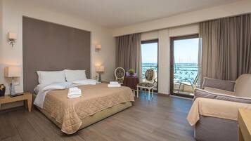 Una habitación de hotel con una cama grande, una zona de estar con un sillón, una pequeña mesa con un jarrón y un balcón con vistas al mar.