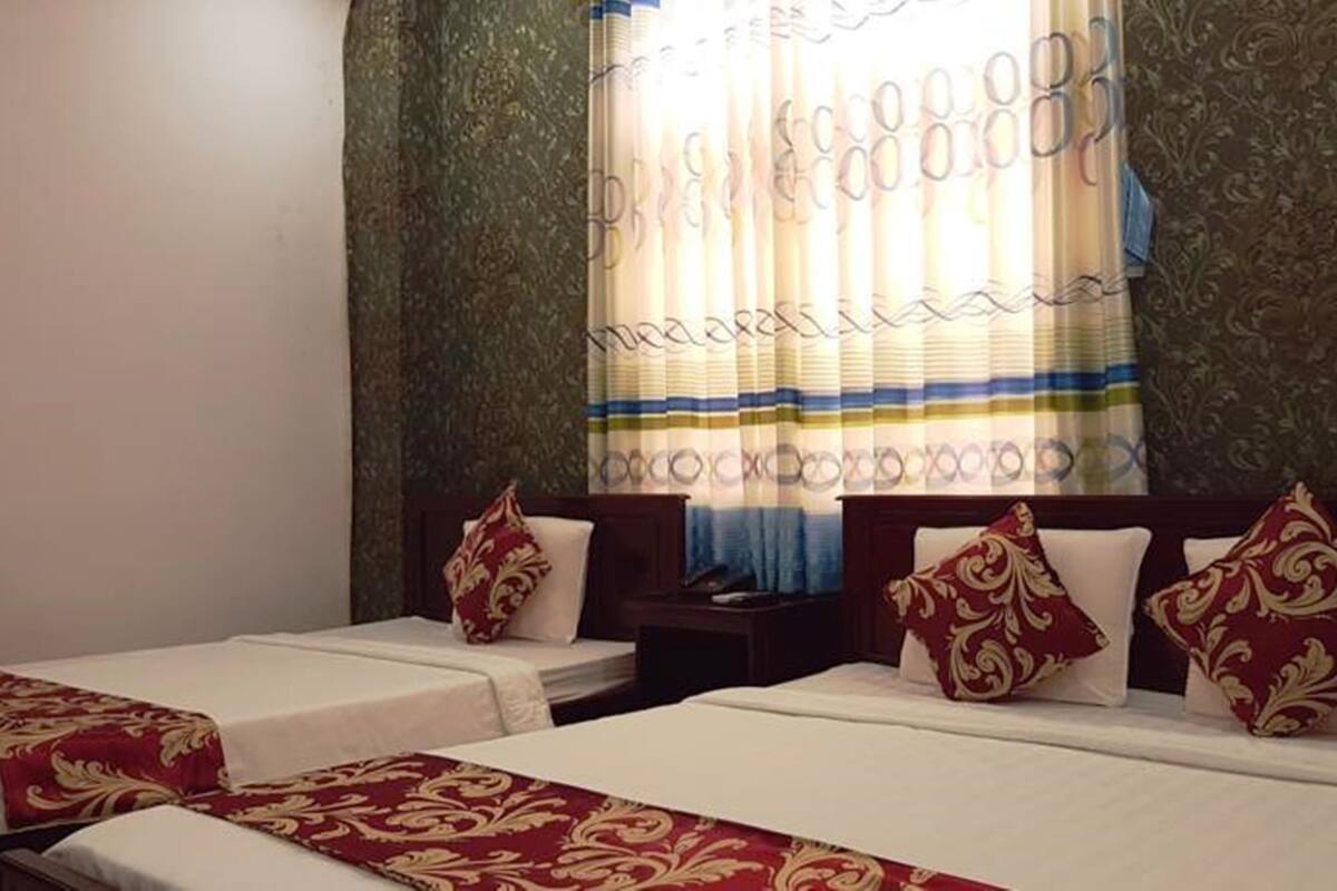 A hotel room with two single beds, each with a red and gold patterned bedspread, and matching pillows. The room has a large window with a patterned curtain, and the walls are adorned with a decorative wallpaper.