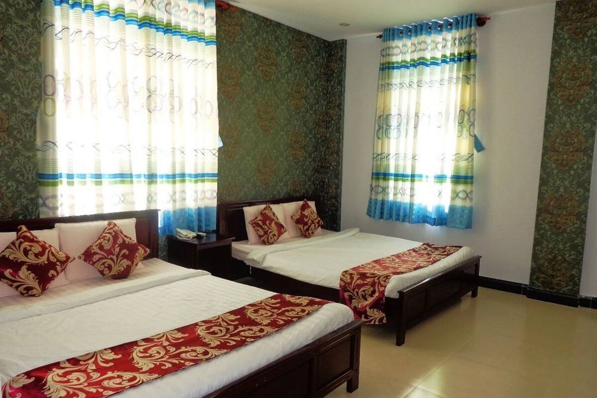 A hotel room with two single beds, each with a red and gold bedspread, and a wooden headboard. The room has patterned wallpaper and curtains with blue and green stripes.
