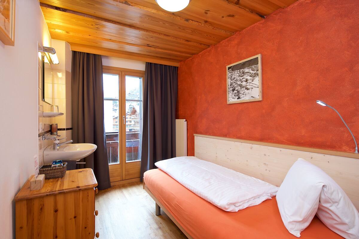 Un dormitorio con cabecera de madera, una cama individual, una mesita de noche con lámpara, una ventana con cortinas y vistas al exterior.