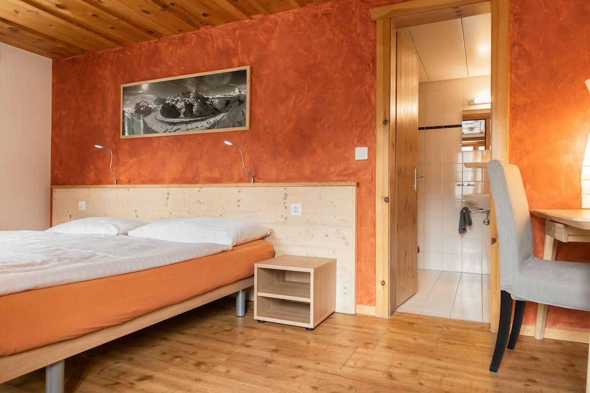 Un dormitorio con cabecera de madera, cama con ropa de cama blanca, mesita de noche pequeña, una silla y una puerta que da al baño.