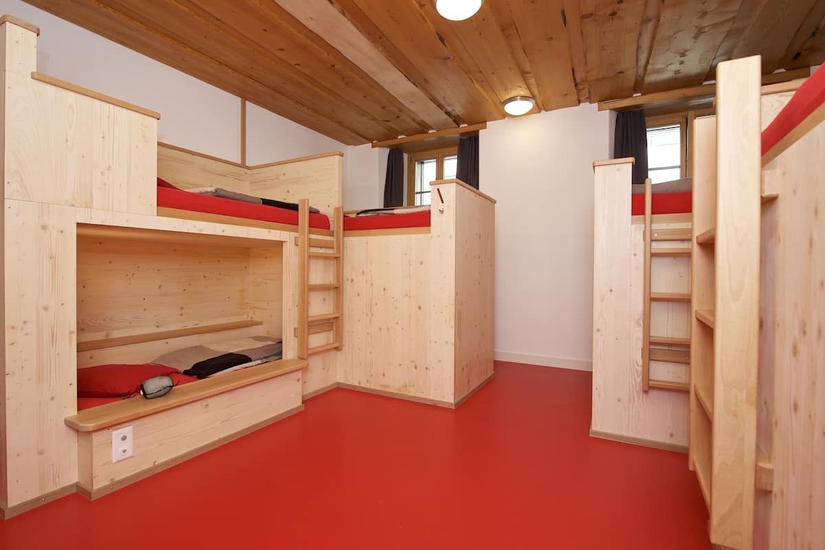 Una habitación con literas, paredes de madera, piso rojo y una ventana con cortinas.