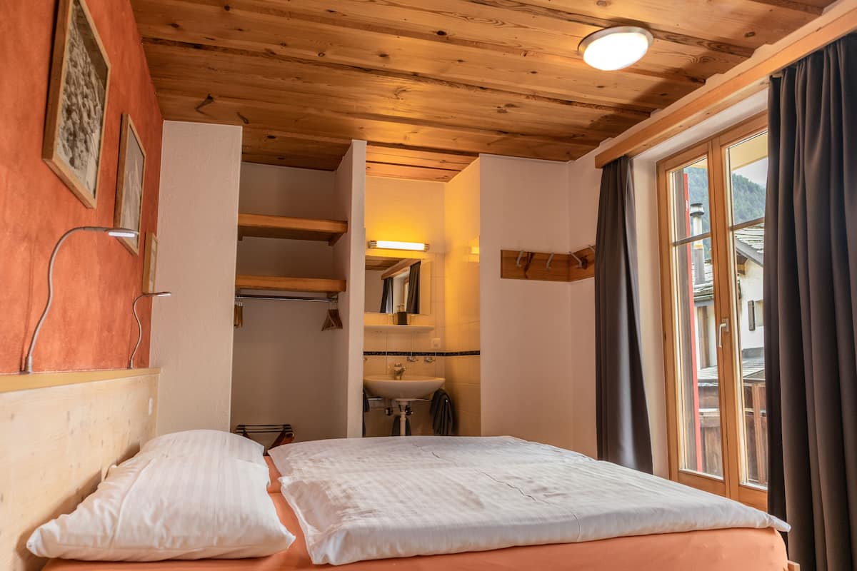 Un dormitorio con techo de madera, una cama con ropa blanca, una ventana con cortinas y un baño visible con lavamanos y espejo.