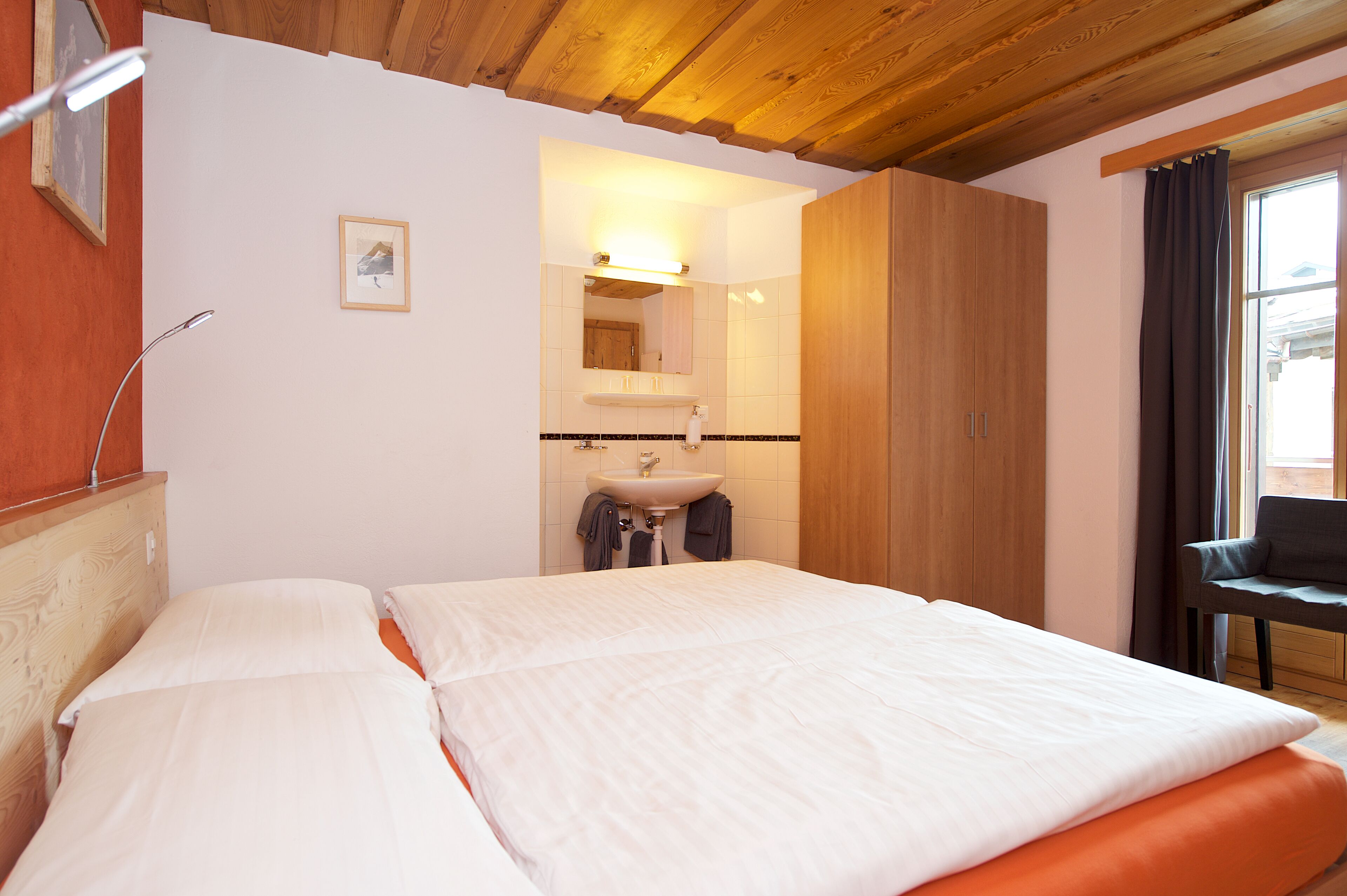 A hotel room with a bed, a wooden ceiling, a bathroom with a sink and mirror, and a window with curtains.