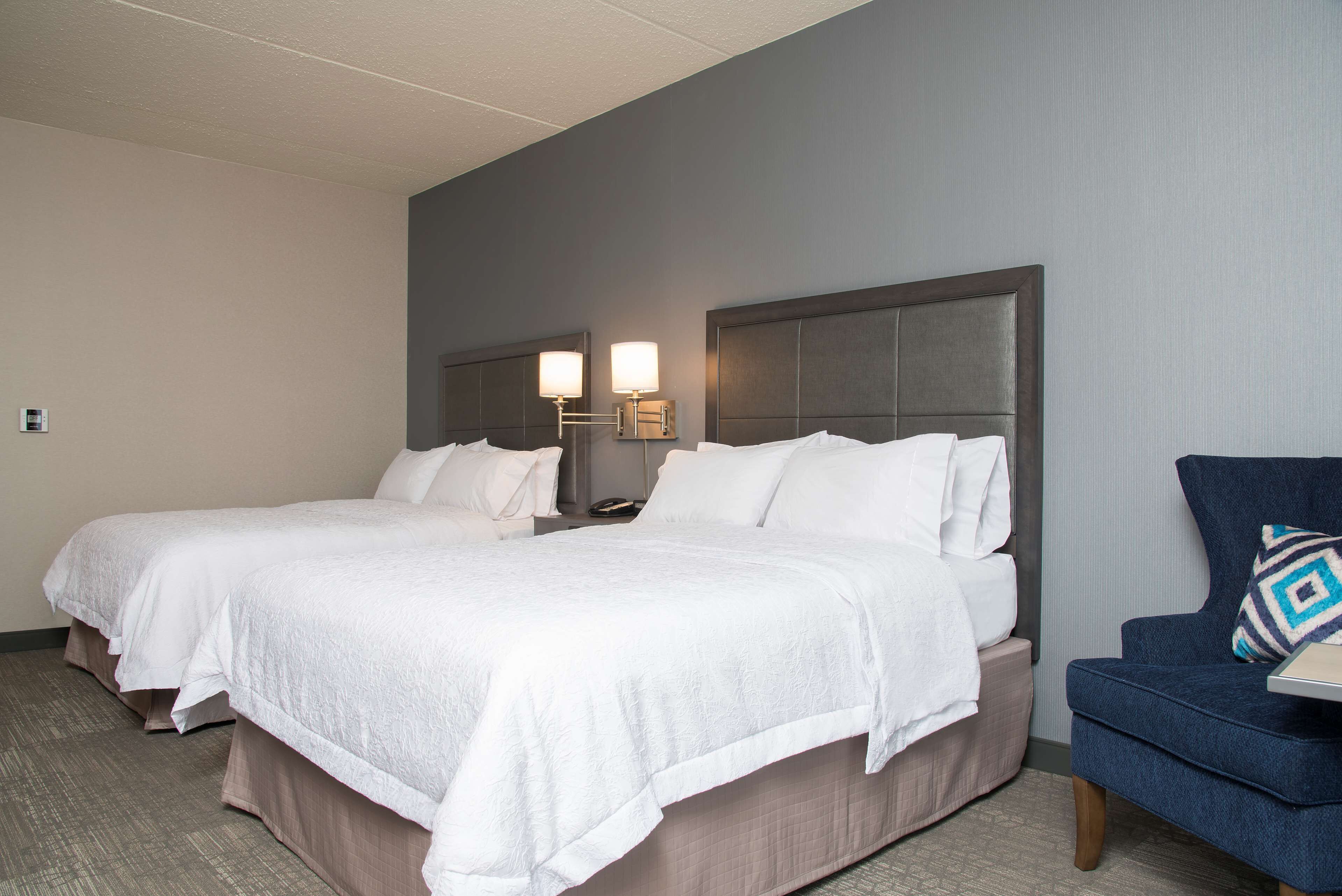 A hotel room with two beds, a nightstand, a lamp, and a chair.