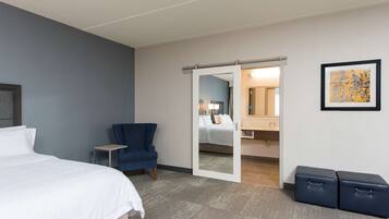 Habitación de hotel con cama, una silla, un escritorio, un espejo y un baño visible a través de una puerta abierta.