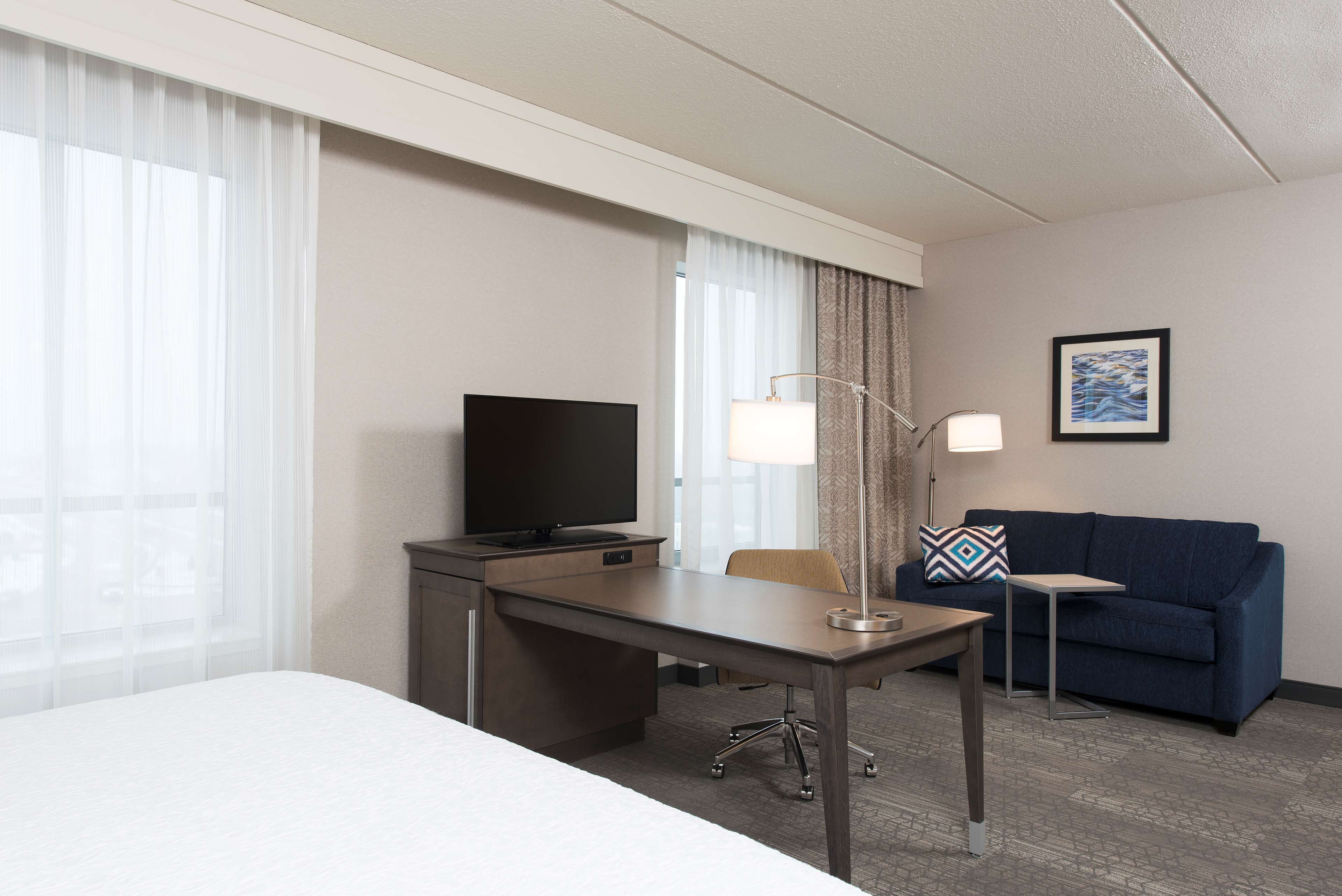 Habitación de hotel con cama, escritorio con televisor, lámpara, sofá e imagen enmarcada en la pared.