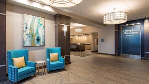 A hotel lobby with two blue armchairs, a large abstract painting, a reception desk, and a Hampton Inn & Suites sign.