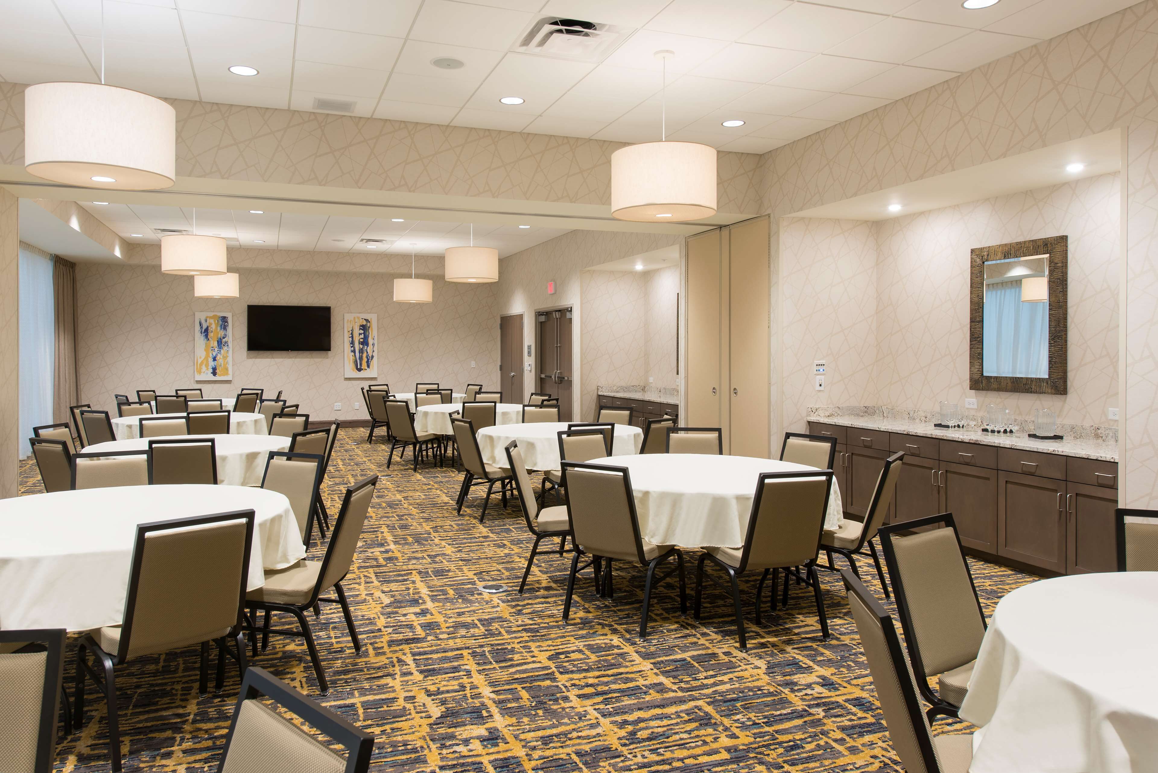 A conference room with multiple tables and chairs, a large flat-screen TV, and a kitchen area with a sink and countertop.