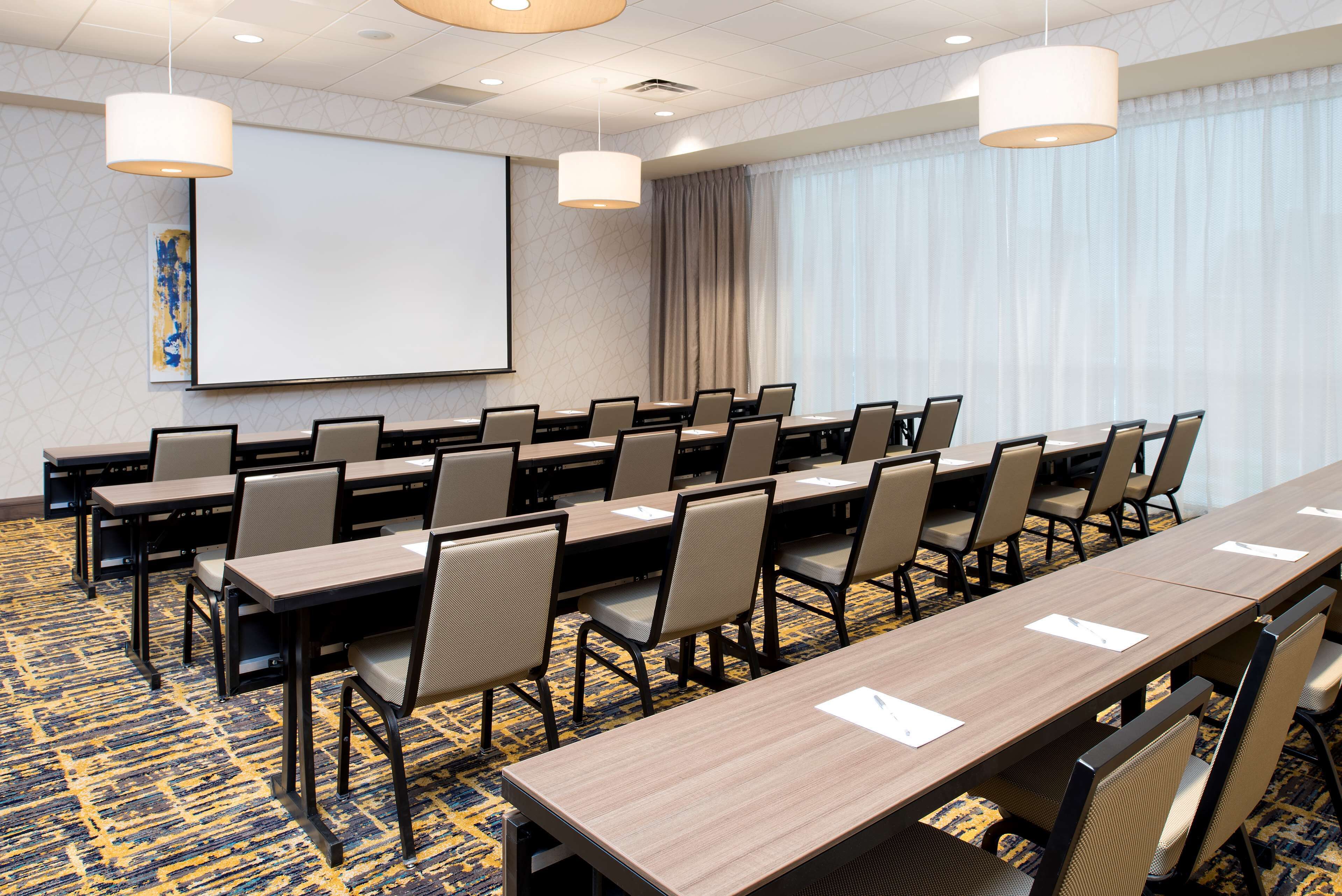 A conference room with multiple tables and chairs, a projector screen, and a large window with curtains.