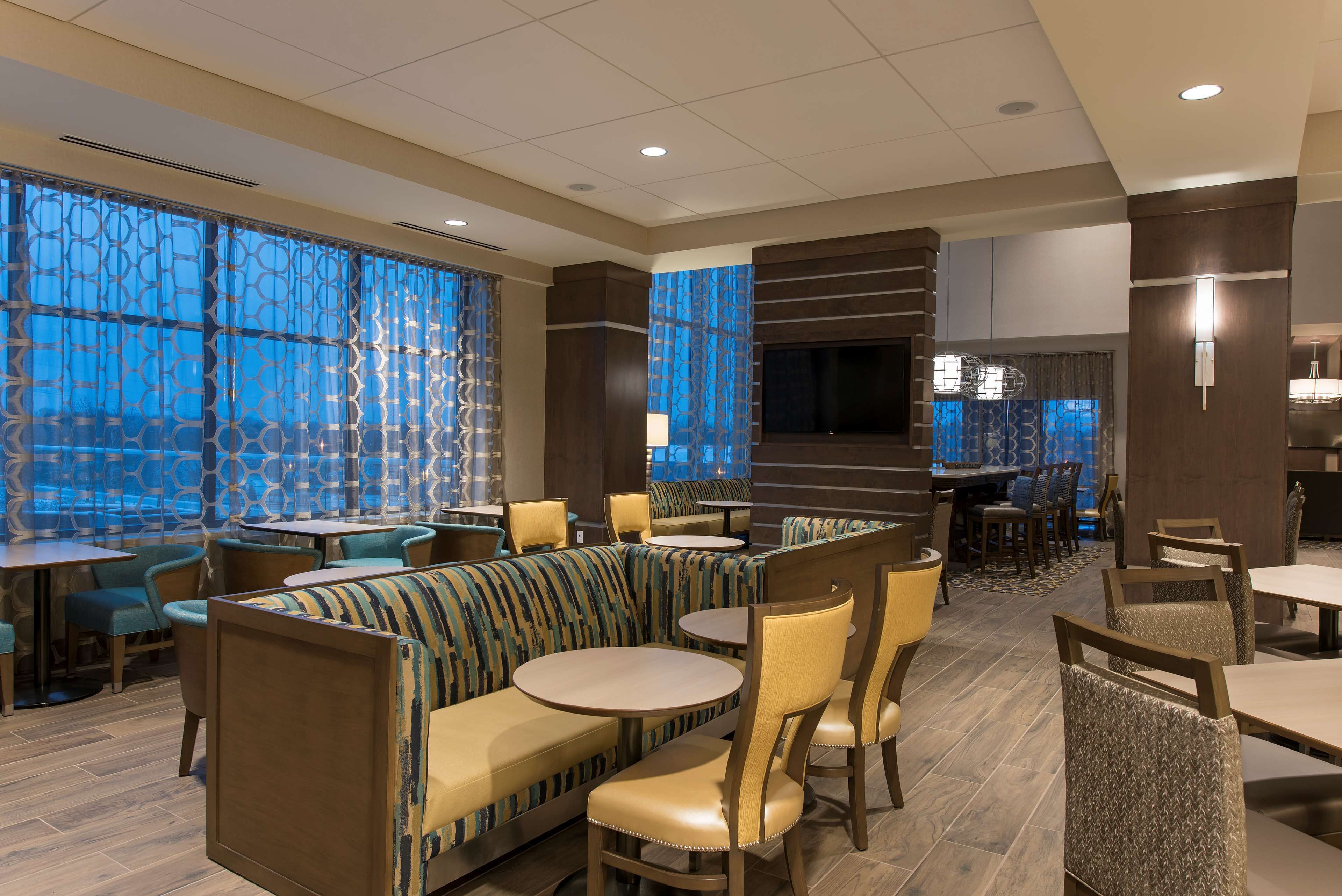 A modern restaurant with booths and round tables, featuring patterned curtains and a large TV screen.