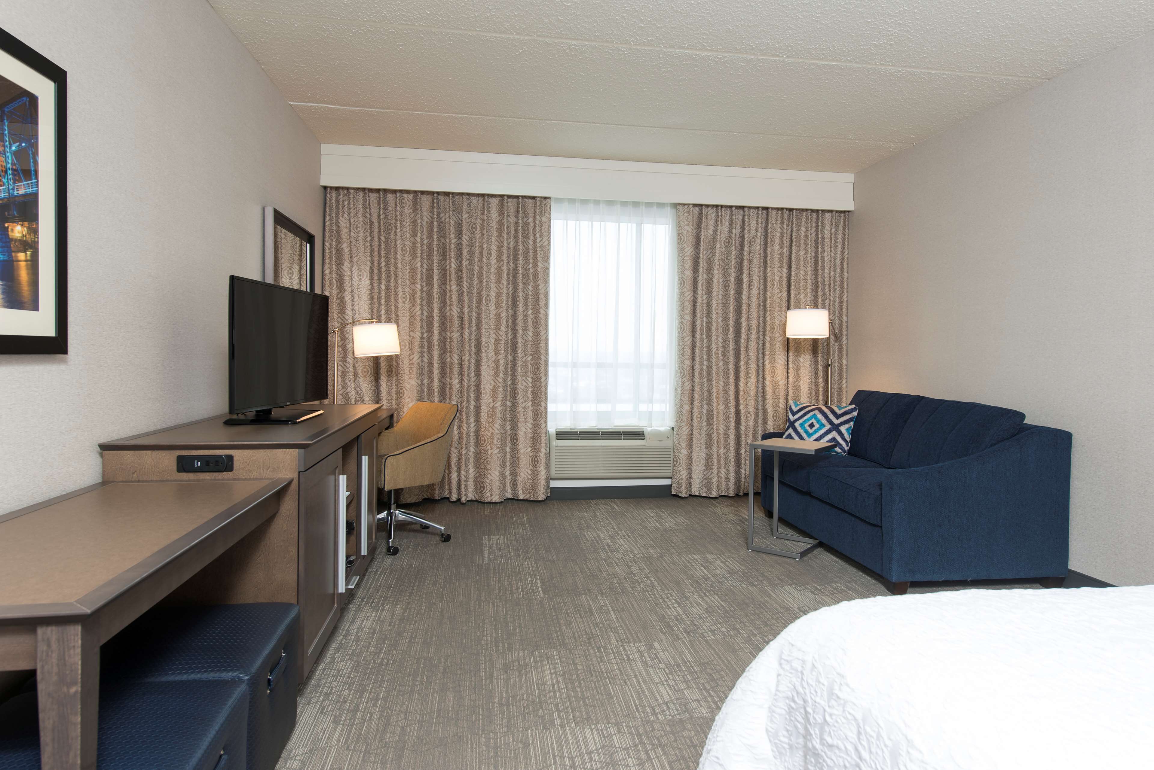 A hotel room with a bed, a desk with a television, a chair, a sofa, and a window with curtains.