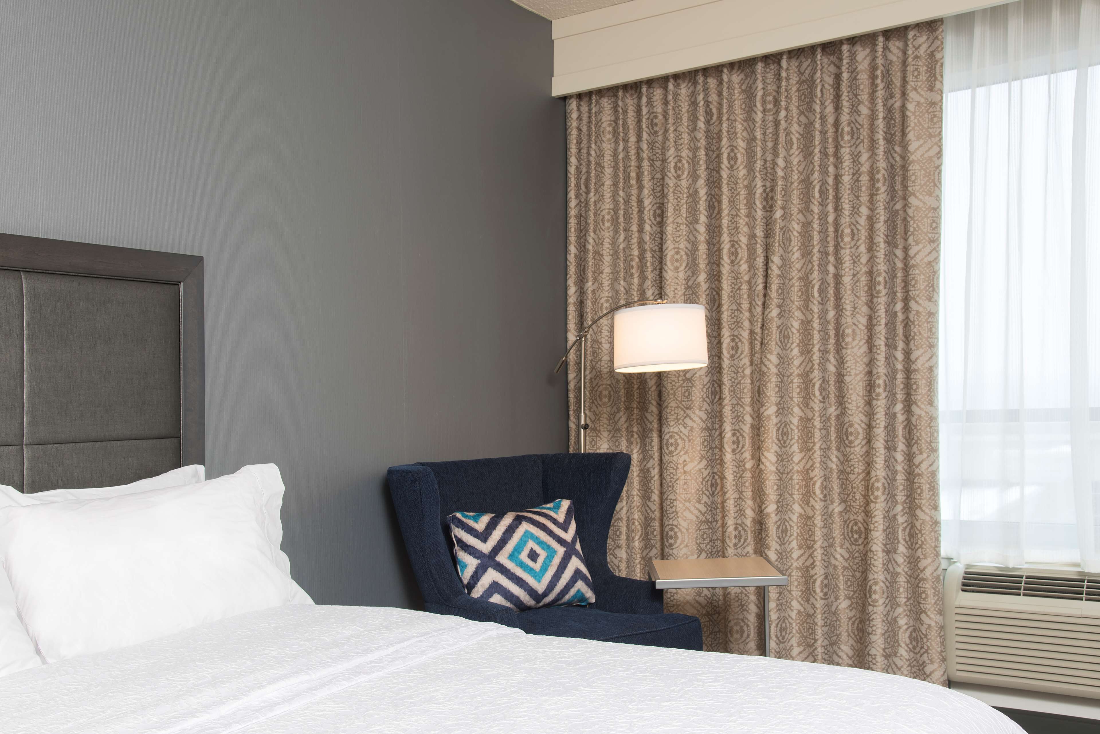 Habitación de hotel con cama, una silla con cojín estampado, mesita de noche y ventana con cortinas.