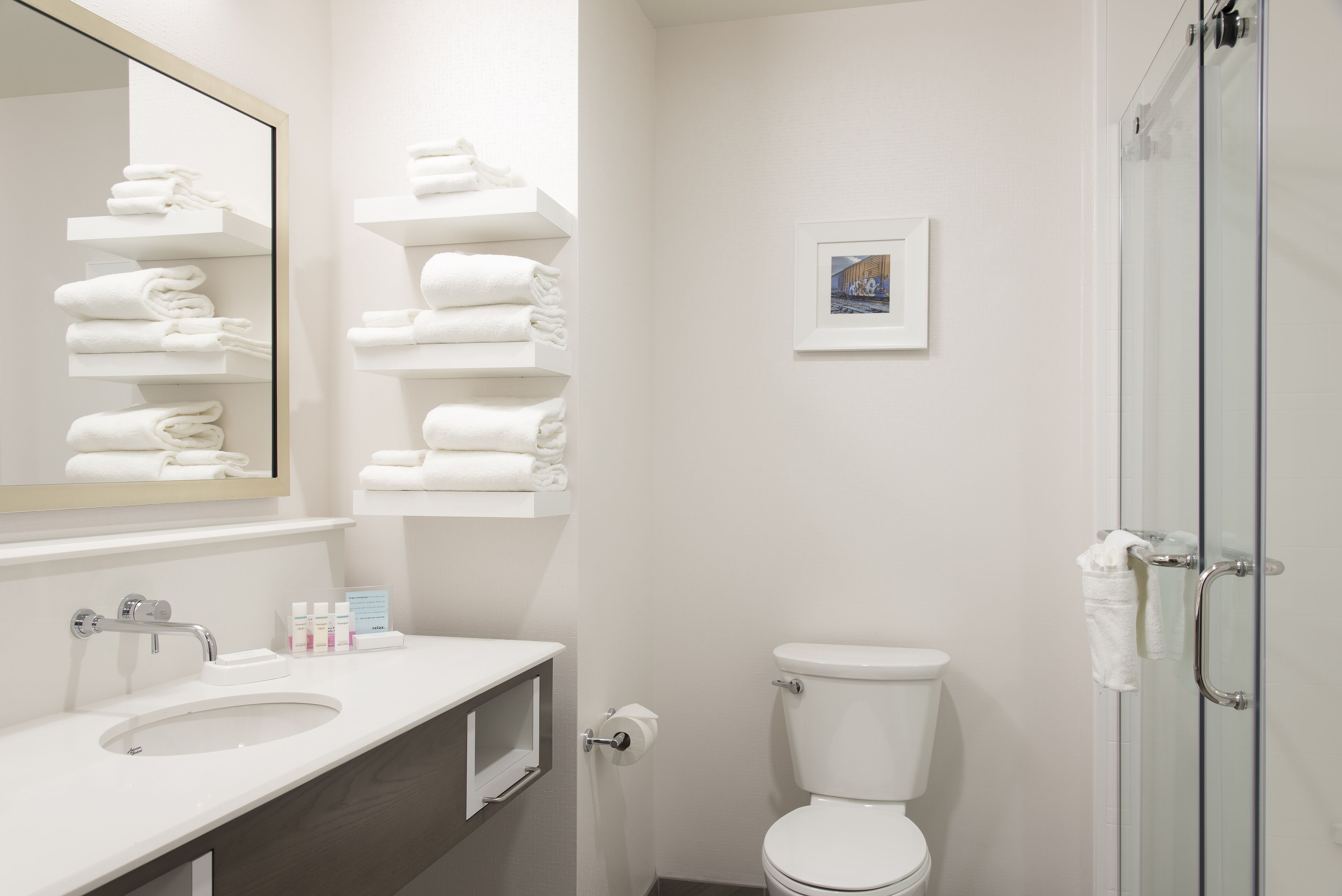 A modern bathroom with a white sink, mirror, and toilet. There are shelves with folded towels and a framed picture on the wall.