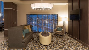 A modern hotel lobby with a patterned carpet, a round ottoman, a sofa, a chair, and a large window with curtains.