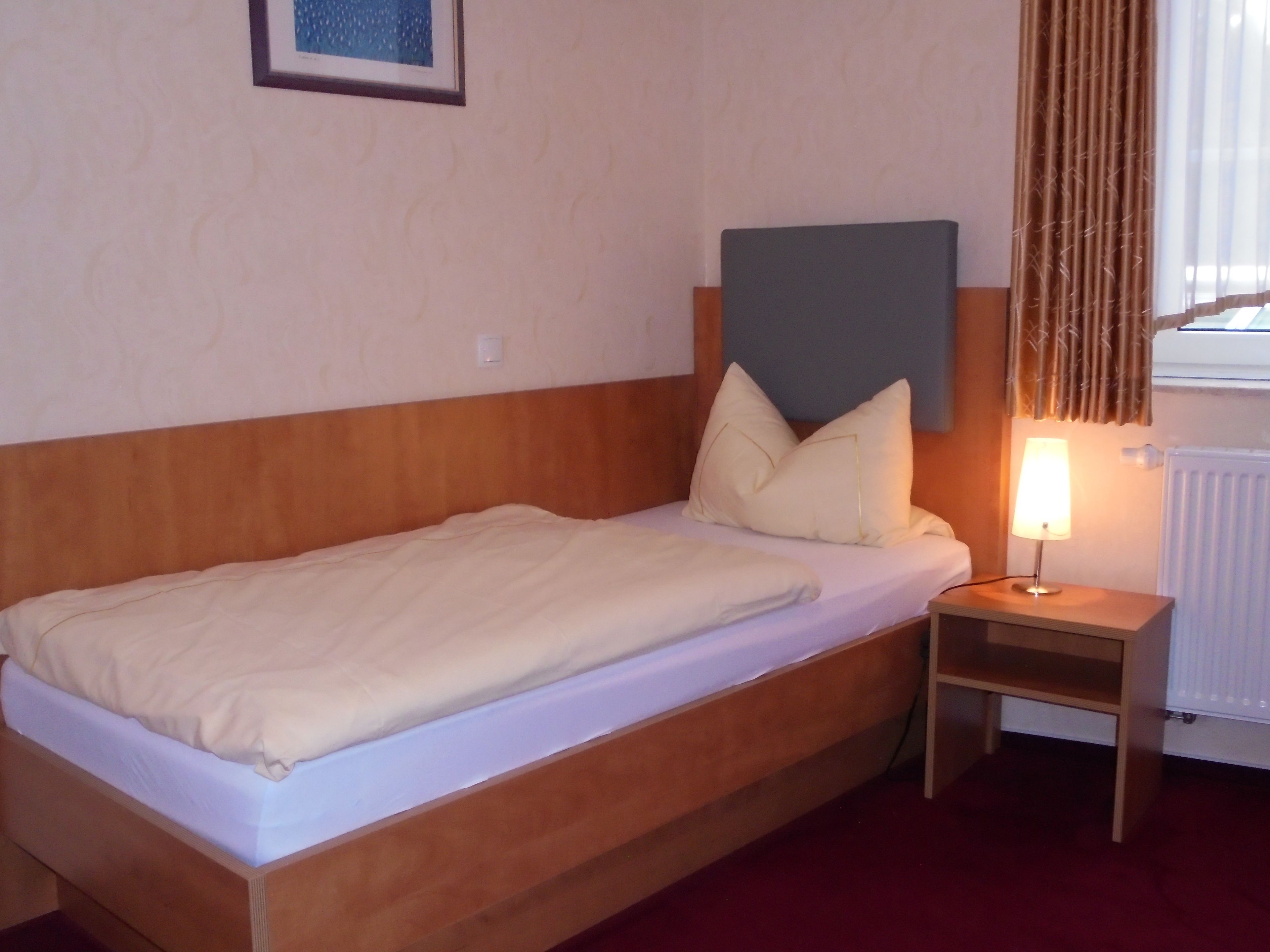 A single bed with a wooden headboard, a bedside table with a lamp, and a window with curtains.
