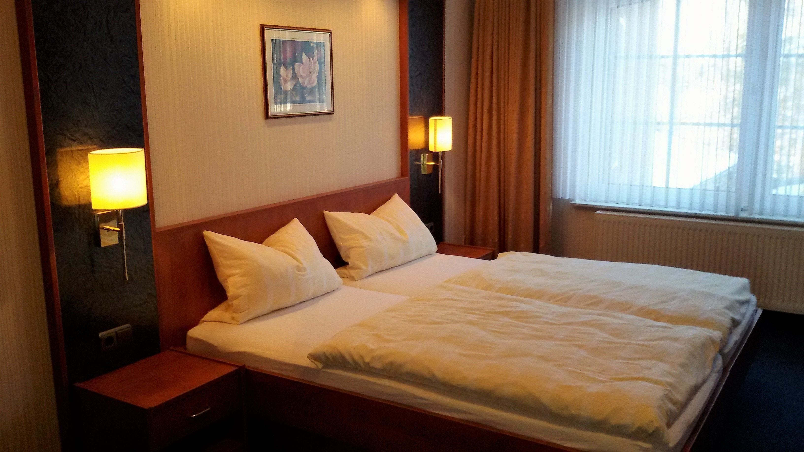 A hotel room with a large bed, two bedside lamps, a framed picture on the wall, and a window with curtains.