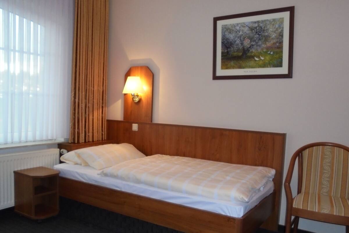 Uma cama de solteiro com cabeceira de madeira, mesa de cabeceira, uma cadeira, uma luminária na parede e um quadro emoldurado.