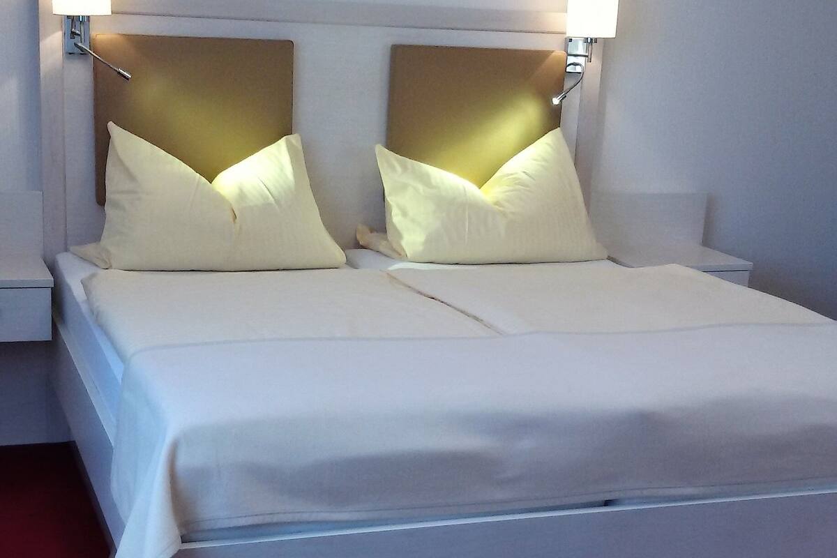 Uma cama bem arrumada com lençóis brancos e duas almofadas, com duas luminárias fixadas na parede e cúpulas brancas.