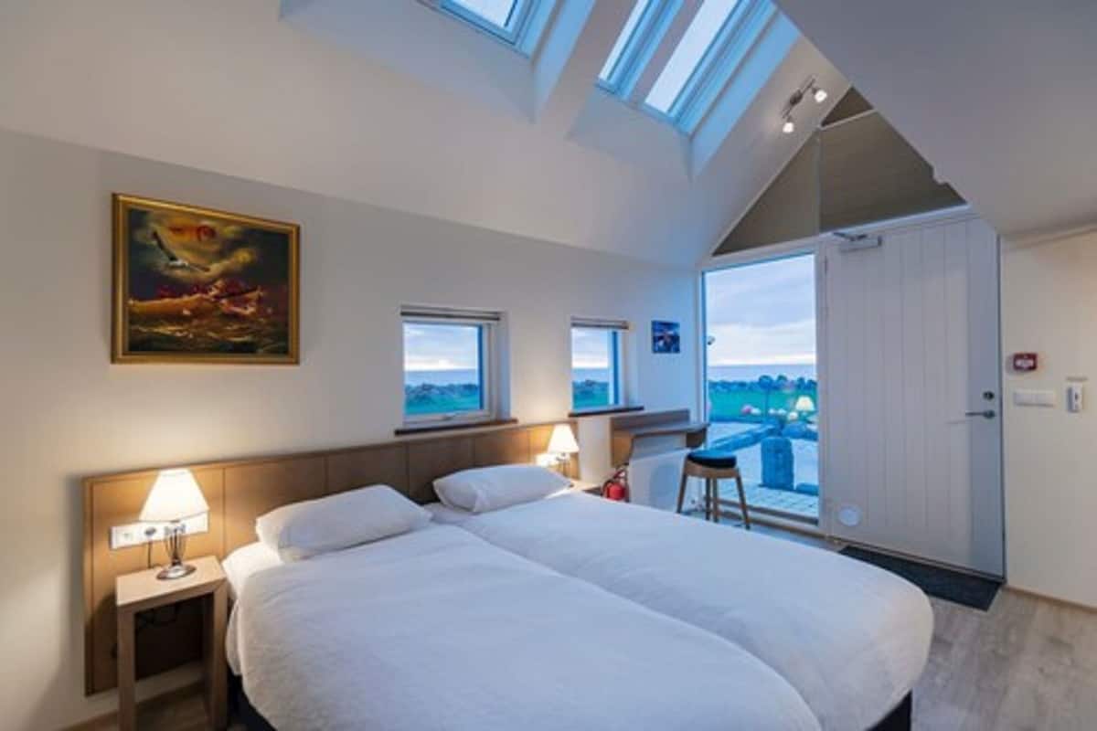 A bedroom with a large bed, two bedside tables with lamps, a painting on the wall, and a view through a door.
