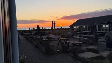 An outdoor seating area with tables and chairs, a building with a sloped roof, and a view of the sunset over the water.