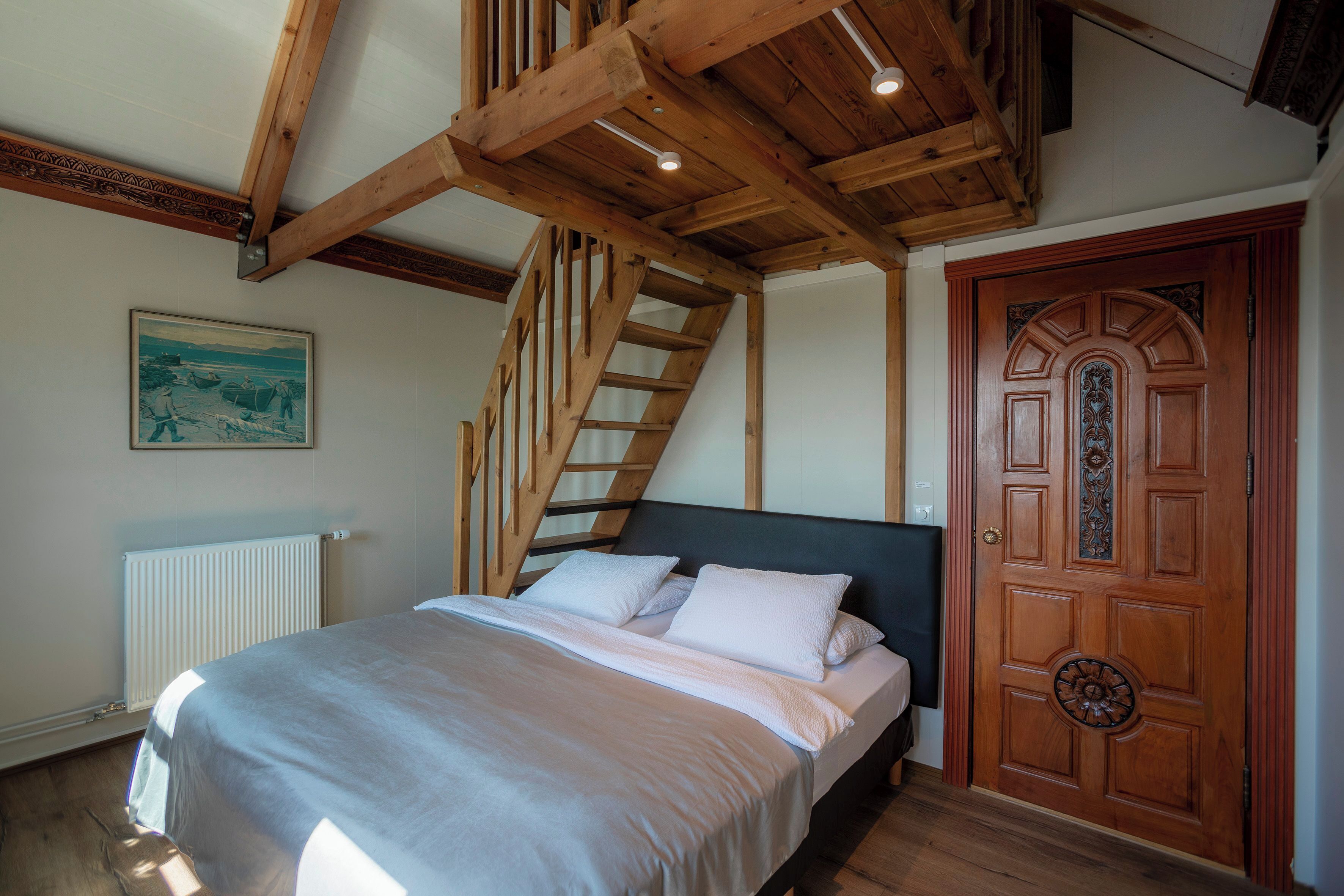 Een slaapkamer met een bed, houten balken in het plafond, een trap, een radiator en een decoratieve deur.
