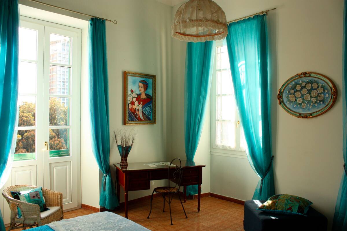A room with a desk, chair, and a bed. There is a window with curtains, a painting on the wall, and a vase with branches.