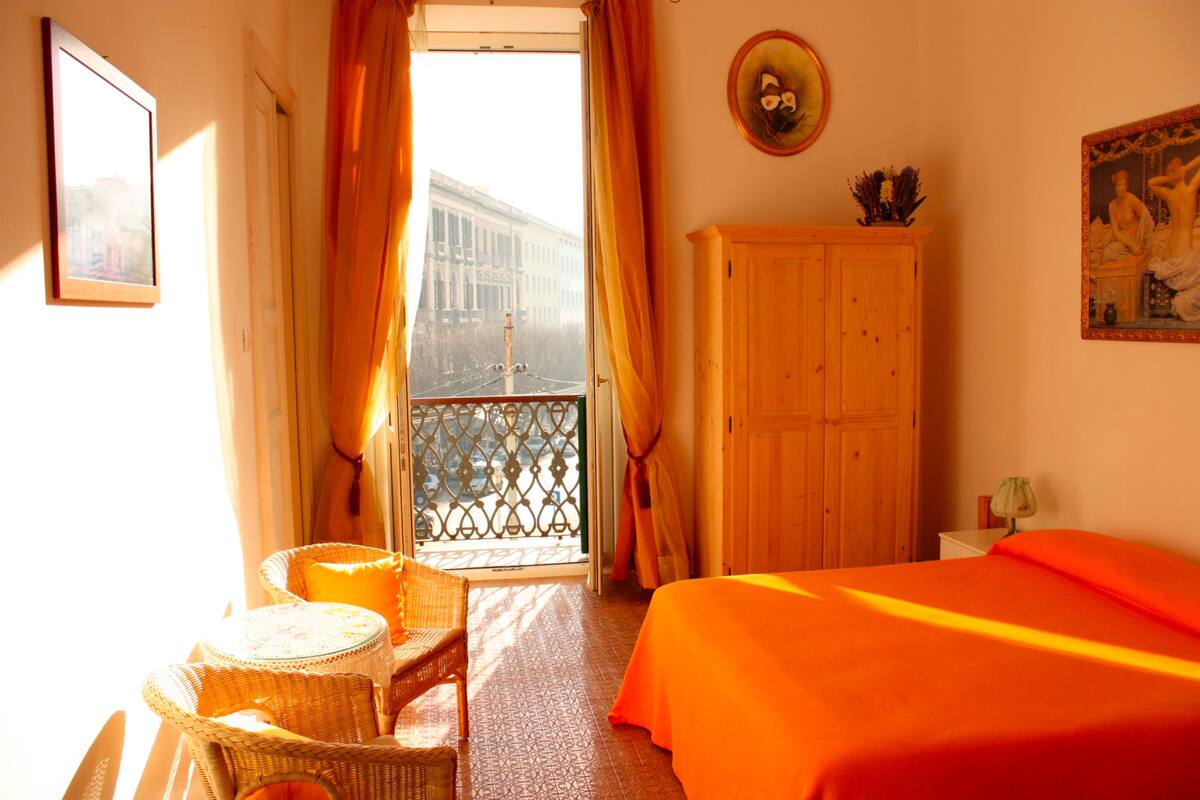 A hotel room with a balcony, a bed with an orange bedspread, a wooden wardrobe, and a wicker chair.
