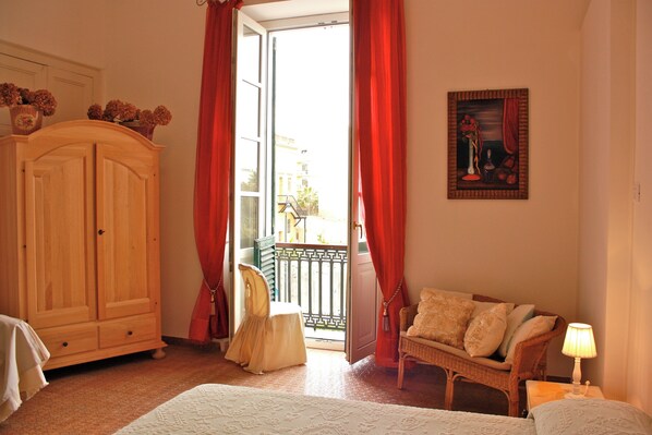 Un dormitorio con un armario de madera, una cama con cubrecama blanco, una silla de mimbre y un cuadro en la pared.
