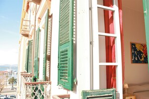Un balcone con persiane verdi, una porta bianca con tenda rossa e un quadro appeso al muro.
