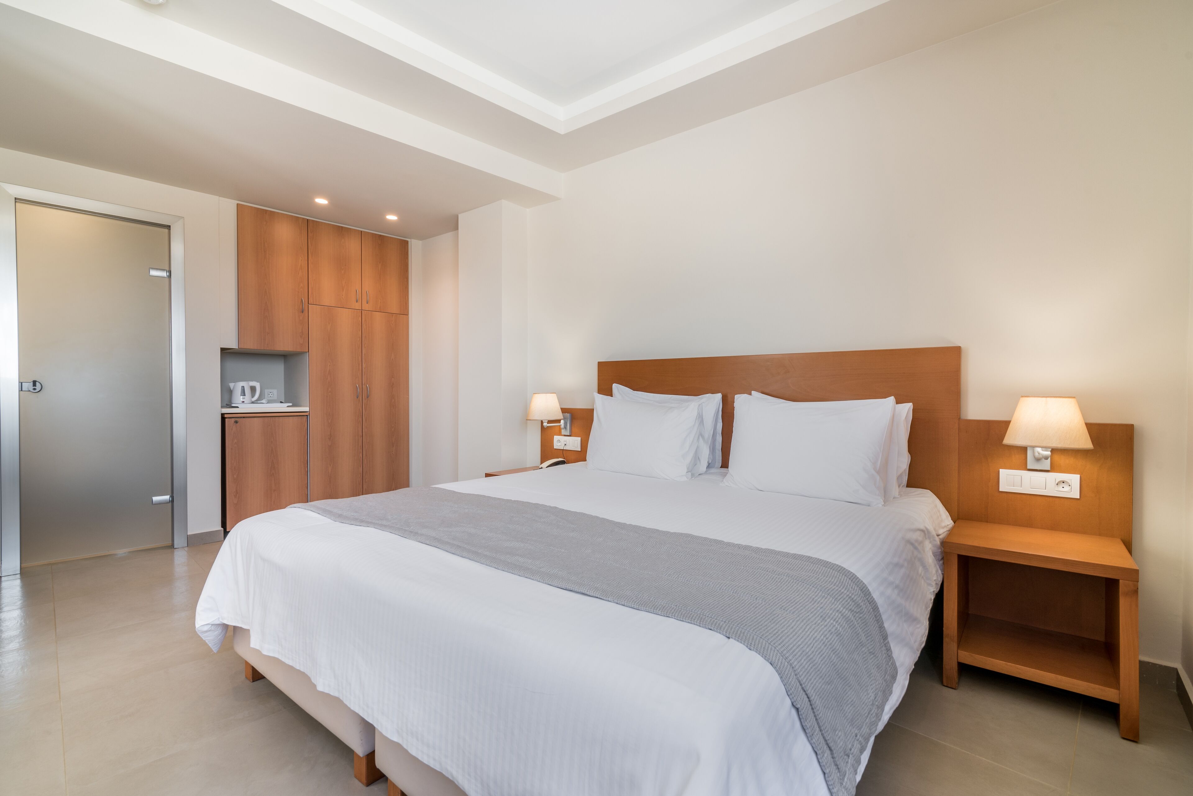 Una habitación de hotel moderna con una cama grande, cabecera de madera, mesitas de noche con lámparas y un armario empotrado.