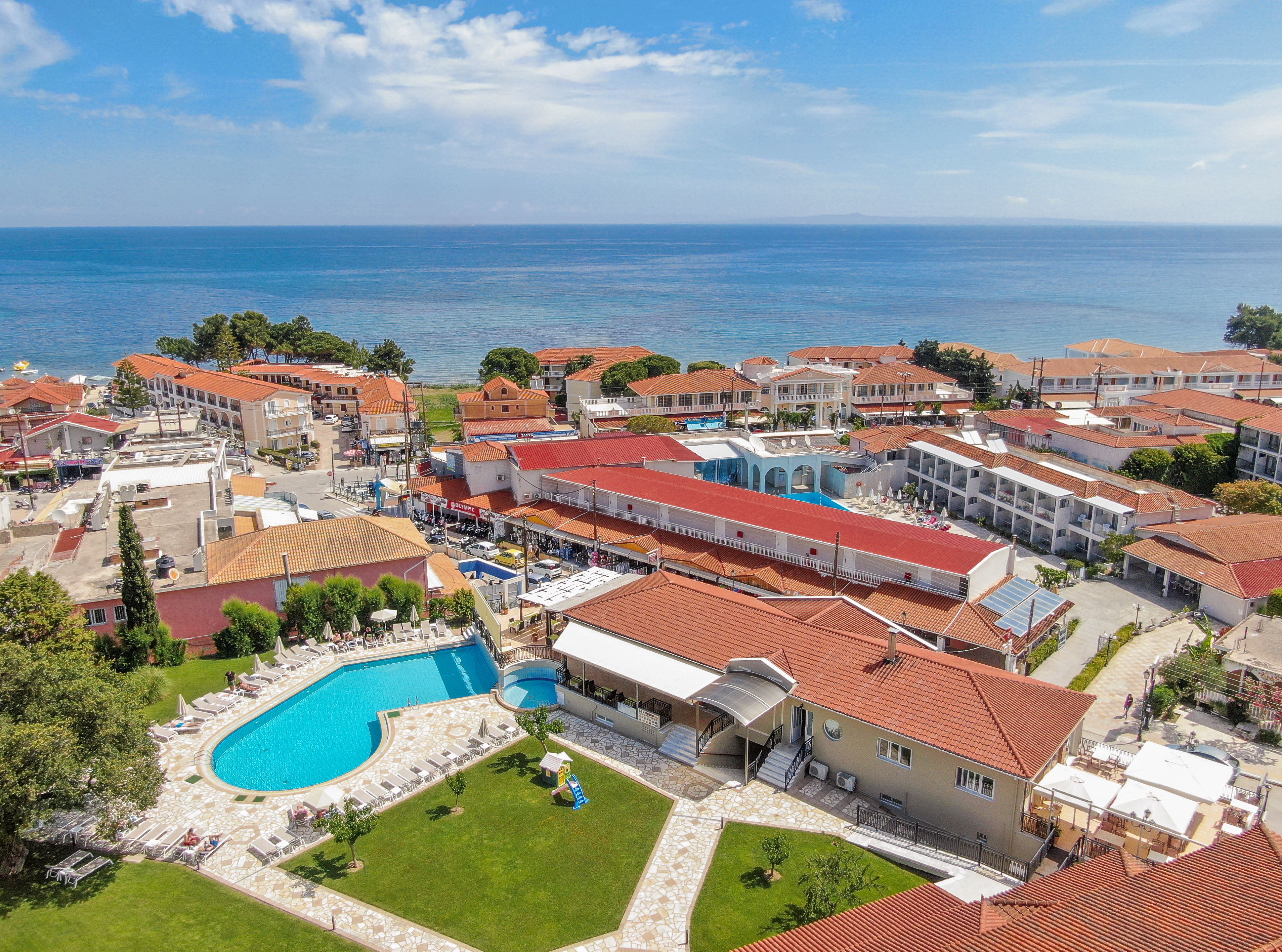A coastal resort with a swimming pool, multiple buildings with red-tiled roofs, and a view of the sea.