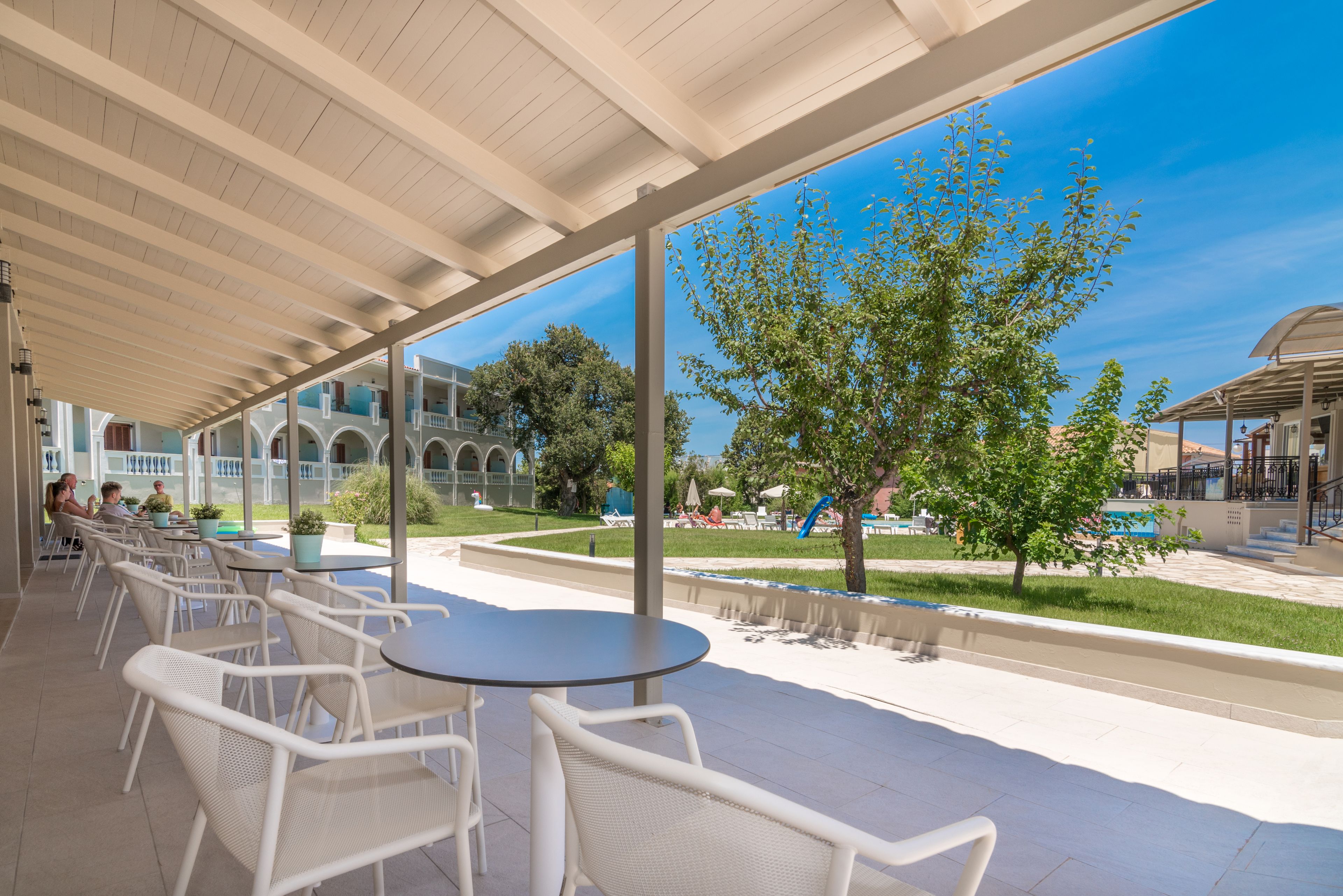 Un patio al aire libre con sillas y mesas de mimbre blanco, un corredor cubierto y vista a una piscina y follaje.