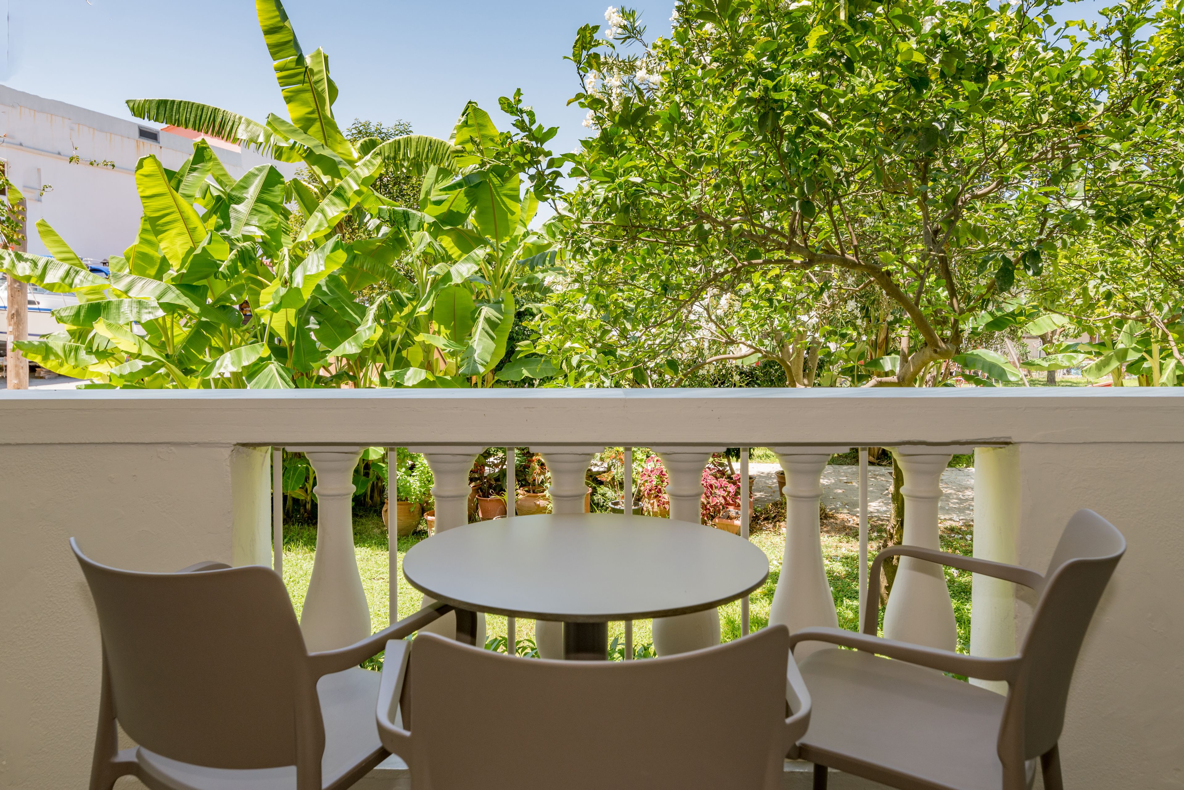A balcony with a round table and four chairs, surrounded by lush greenery and a white fence.