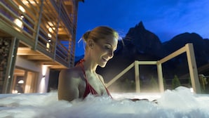 A person in a hot tub with a mountainous backdrop and a building with balconies. - Hotel Arvina (Castelrotto)