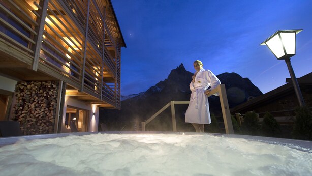A person in a white robe standing by a hot tub with a mountain backdrop. - Hotel Arvina (Castelrotto)