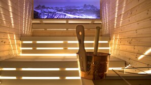 A sauna room with wooden walls, a television screen displaying a snowy mountain landscape, and a wooden bench with a bucket and spoon. - Hotel Arvina (Castelrotto)