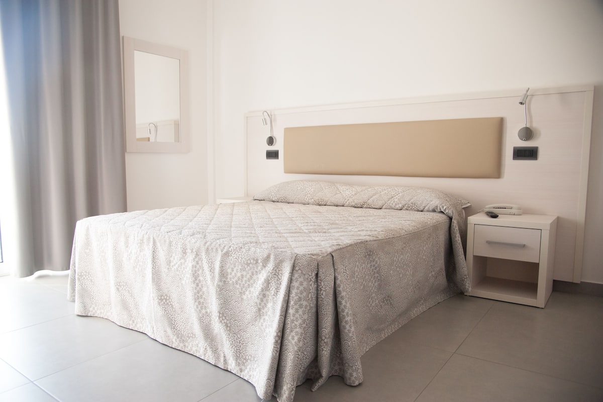 A neatly made bed with a quilted cover, a bedside table with a lamp, and a wall-mounted mirror.