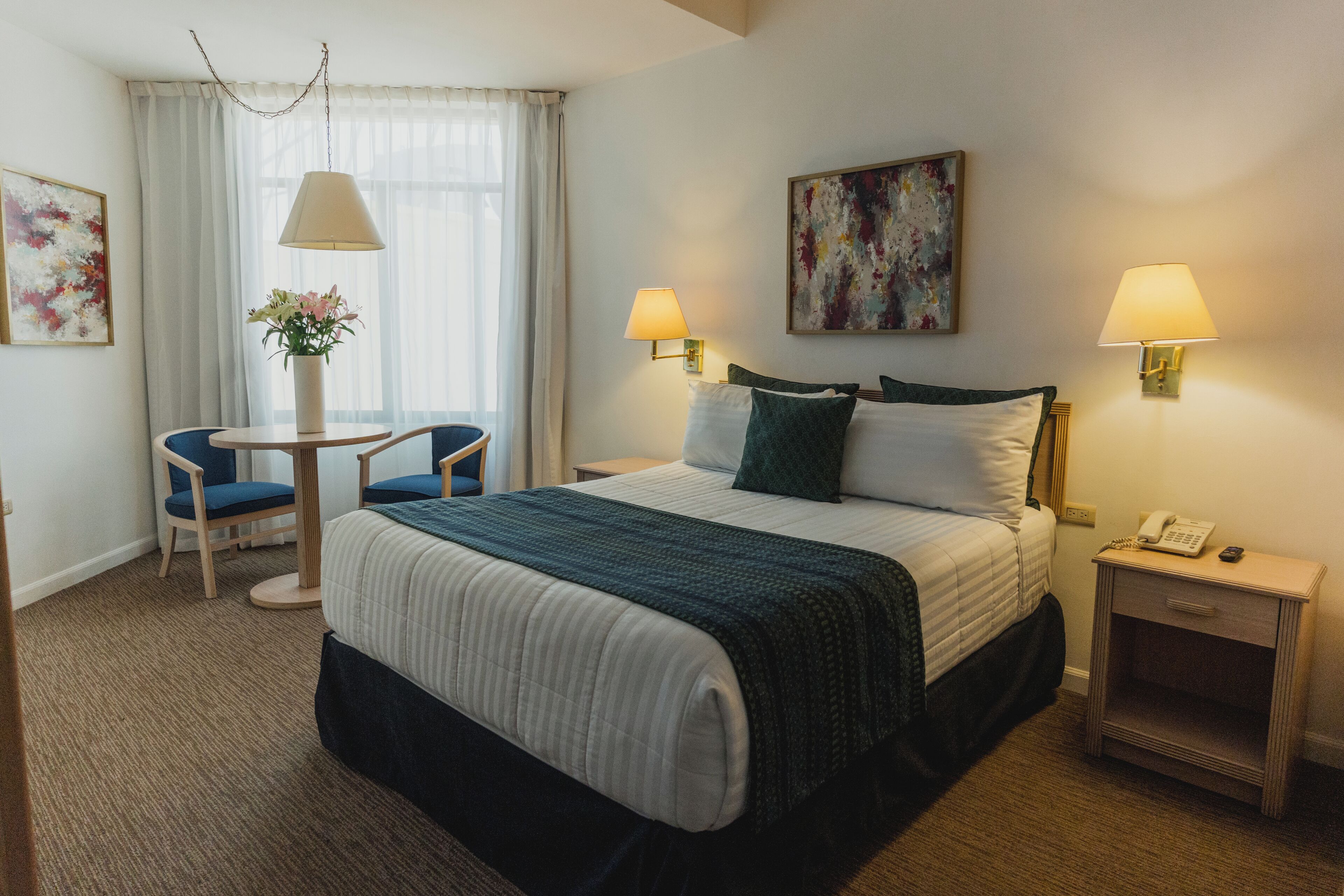 Habitación de hotel con una cama, dos lámparas de noche, un escritorio con una silla, un florero y un cuadro en la pared.