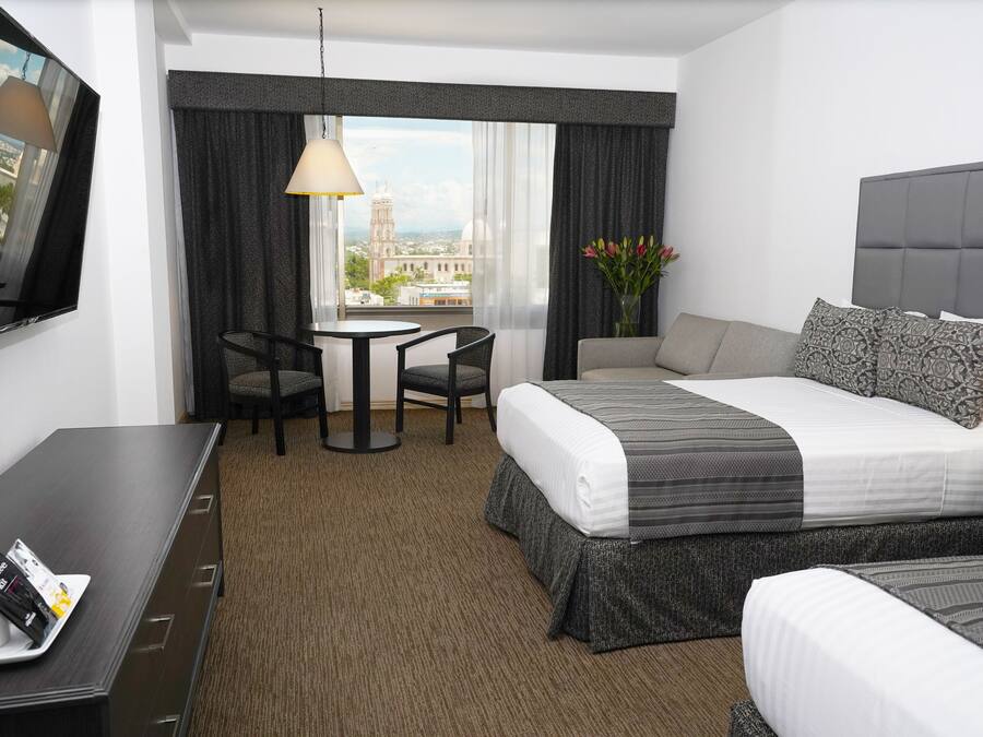 A hotel room with a bed, a desk, a chair, a small table, and a view of a cityscape.
