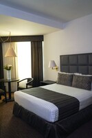 Habitación de hotel con una cama grande, una mesita de noche, una silla, una lámpara y un ventanal con cortinas.