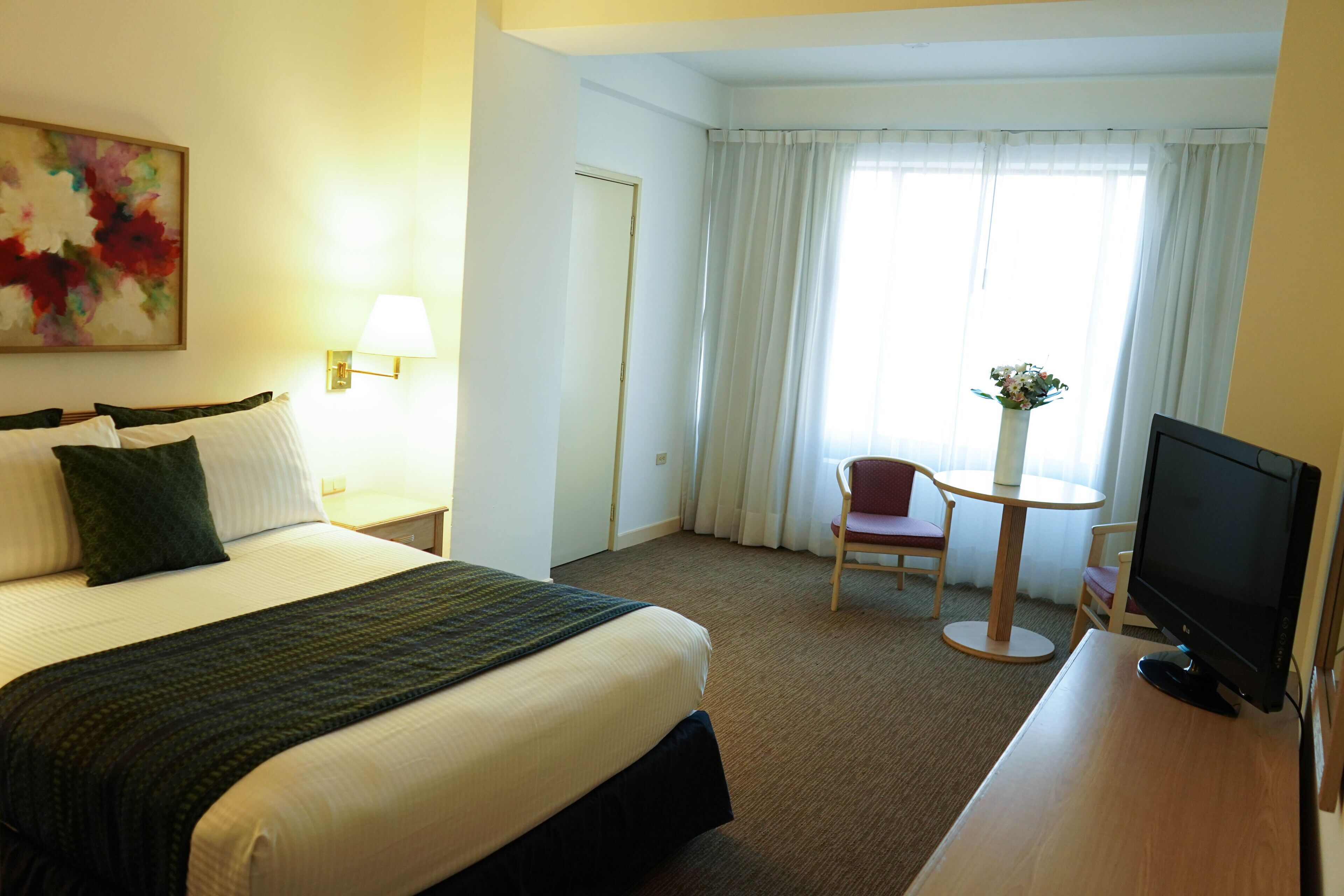 A hotel room with a bed, a desk with a TV, a chair, a small table, and a window with curtains.
