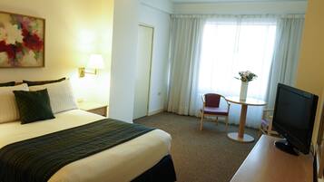 A hotel room with a bed, a desk with a TV, a chair, a small table, and a window with curtains.