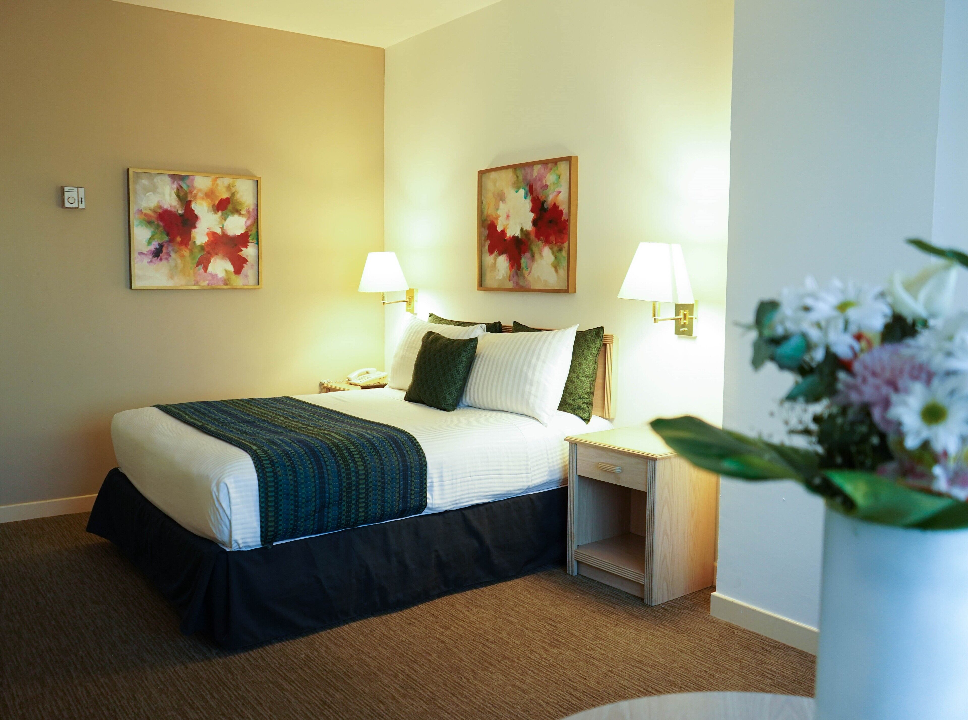 Una habitación de hotel con una cama, dos mesitas de noche, una lámpara y dos cuadros con motivos florales en las paredes.