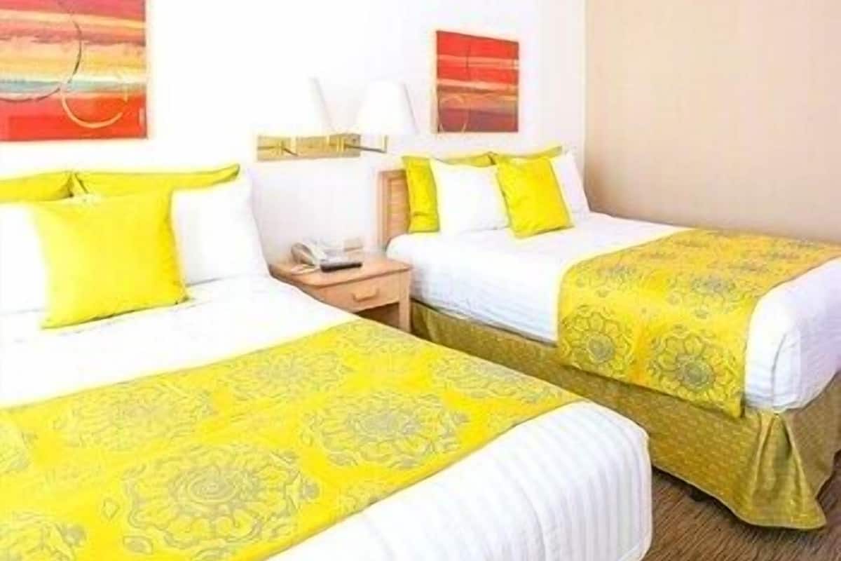 Two twin beds with yellow and white bedding, a bedside table with a lamp, and artwork on the walls.