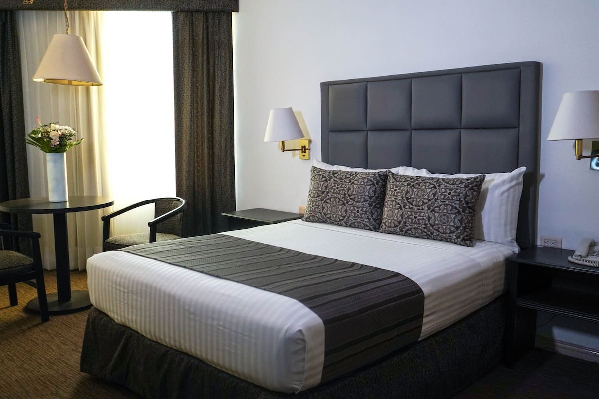 A neatly arranged hotel room with a large bed, two bedside lamps, a small table with a chair, and a vase with flowers.