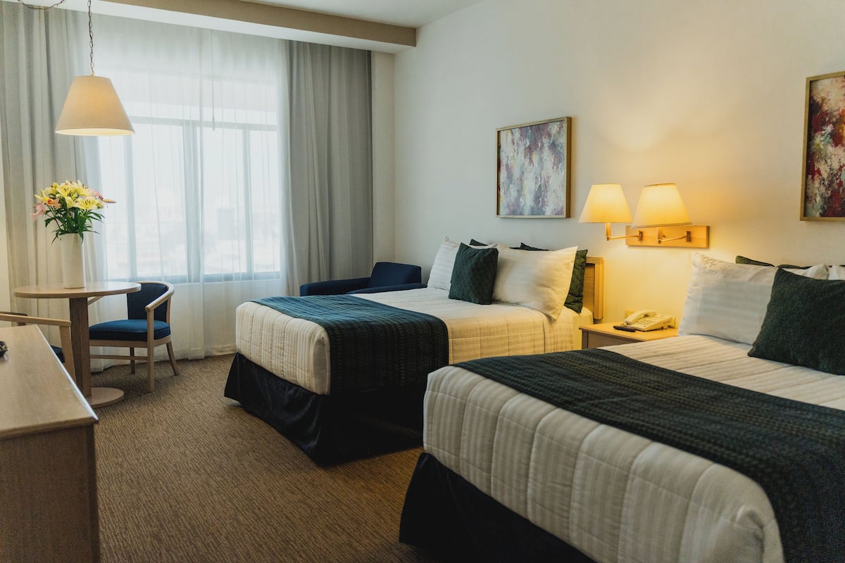 A hotel room with two beds, a desk, a chair, a lamp, and a painting on the wall.