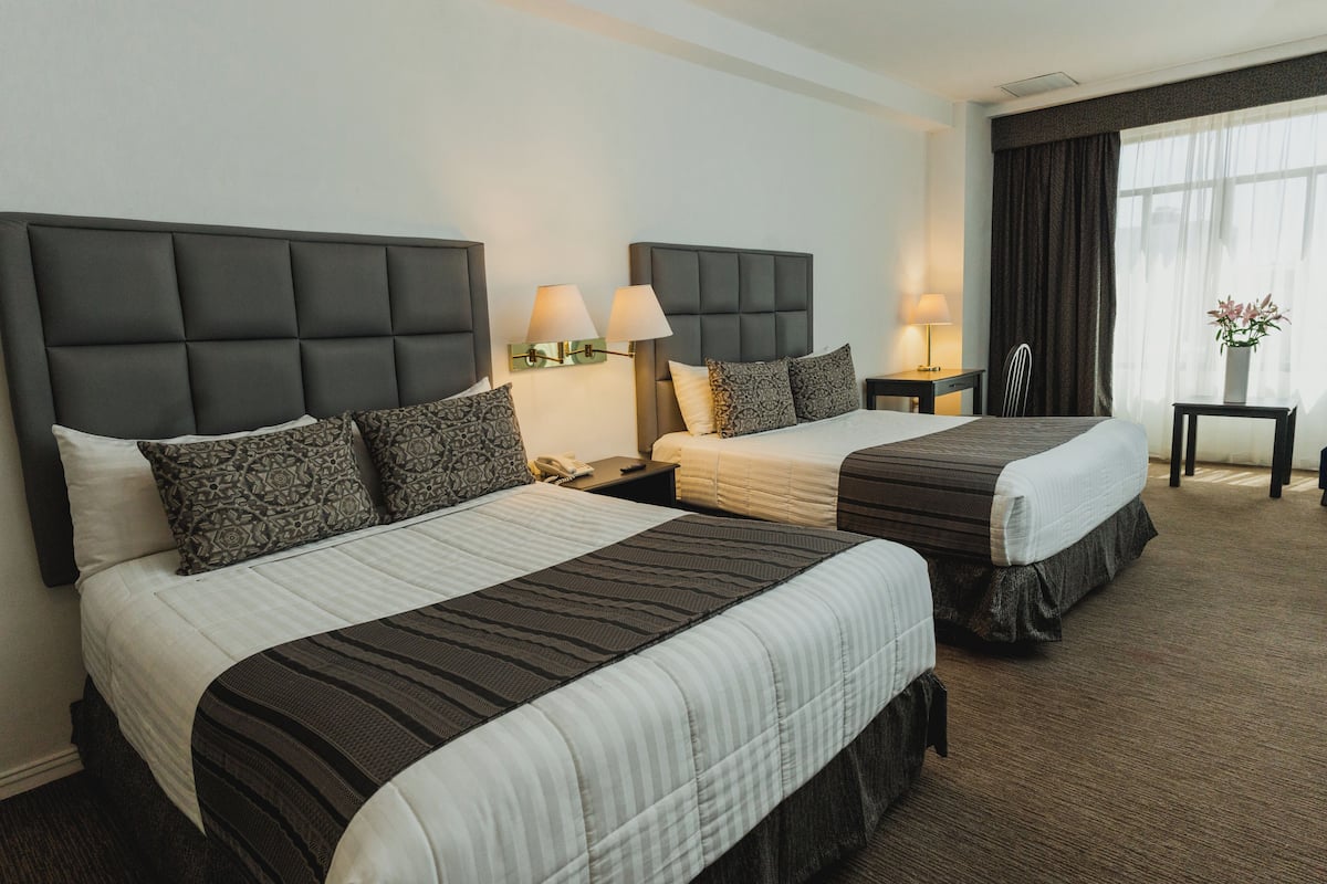 A hotel room with two beds, each with a headboard, bedside tables, lamps, and a window with curtains.